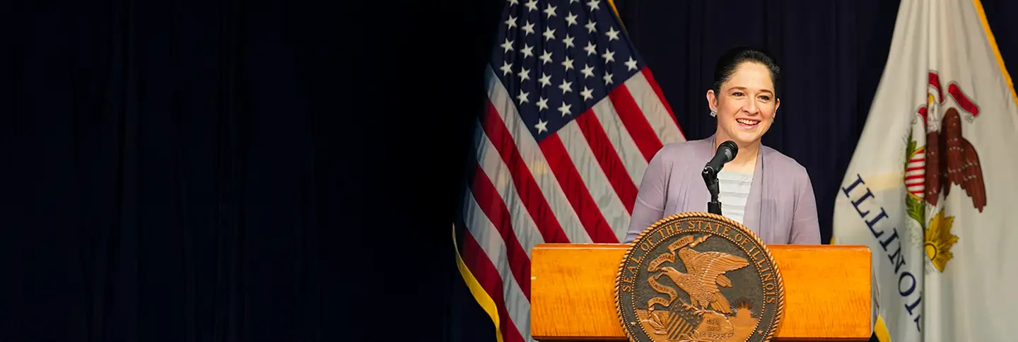 Susana Mendoza speaking at a podium with the American flag and the State of Illinois flag behind her. The Seal of the State of Illinois is on the front of the podium