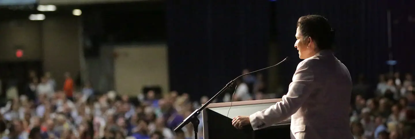 photo of Susana Mendoza wearing a white suit standing at a podium giving a speech to a large crowd