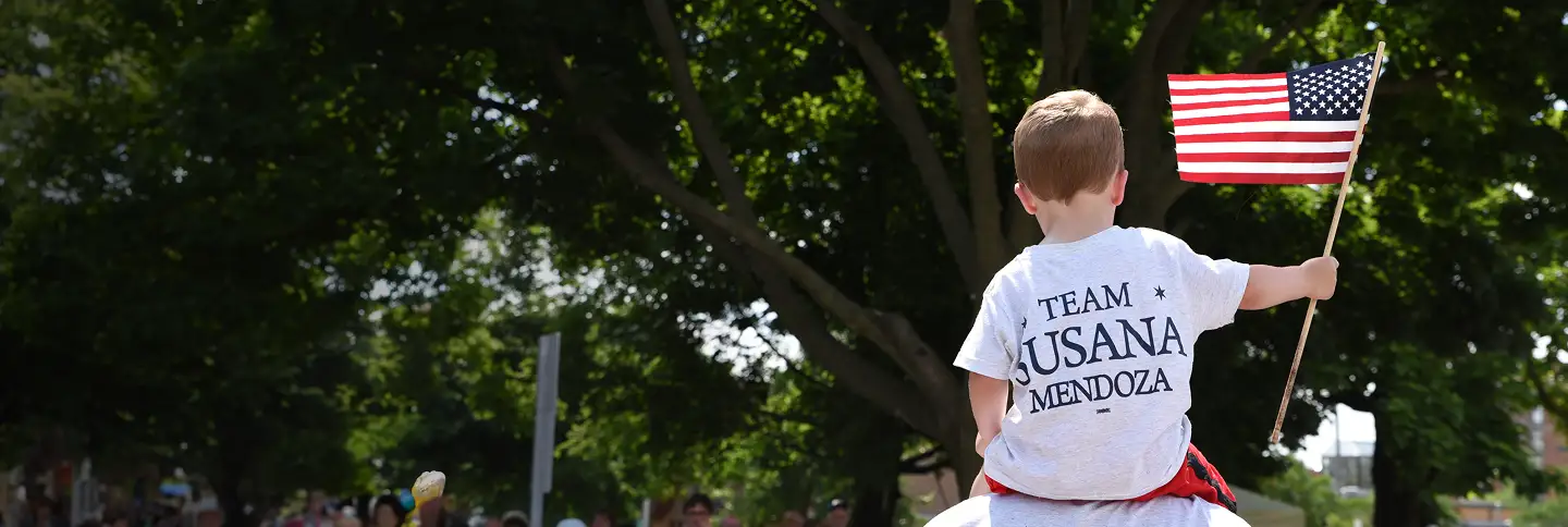 Photo of the backside of a boy holding an American flag, wearing a Team Susana Mendoza shirt and sitting on top of someone's shoulders with green trees in the background